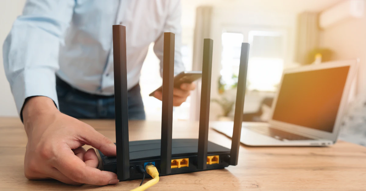Person adjusting a black WiFi router with antennas on a desk next to a laptop