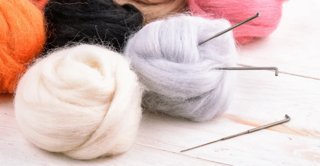 Balls of soft wool yarn and raw wool fibers in pink, gray, and orange with knitting needles