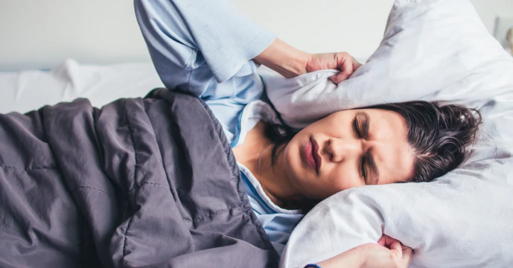 Man awake in bed with a pillow over his head, unable to sleep